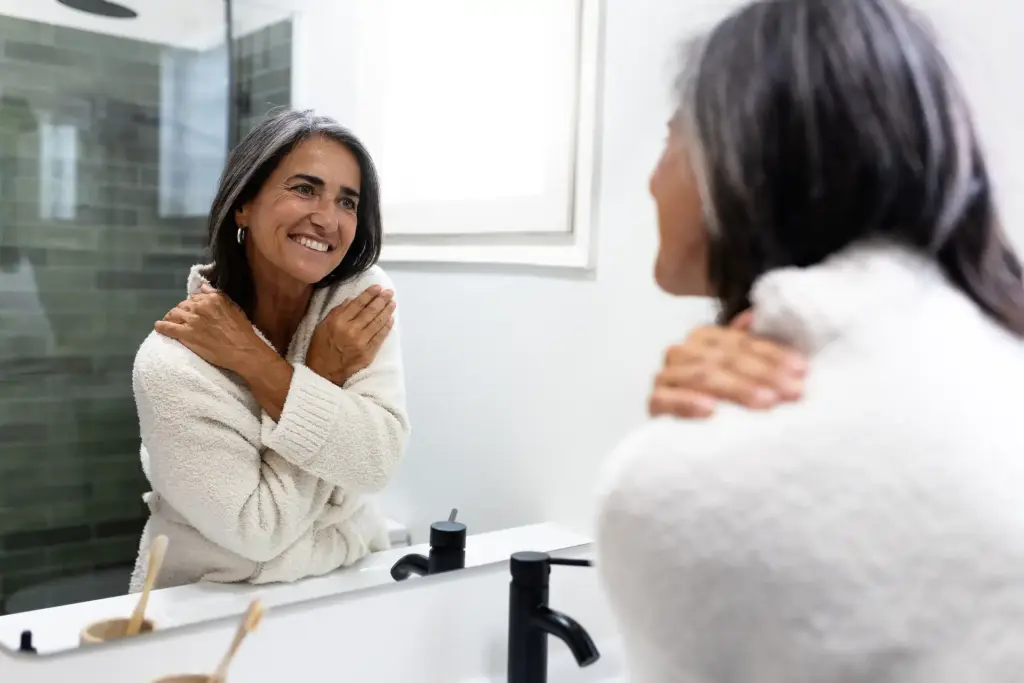 Dona adulta somrient al mirall del bany, mostrant els resultats harmoniosos d'una pell cuidada i rejovenida.